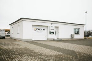 Einfache, weiße Gewerbeimmobilie mit Flachdach, Garagentor und Eingangstür, auf dem Garagentor und neben der Tür das Logo und Schriftzug „DETA Heizung - Service - Lüftungstechnik“, davor gepflasterter Parkplatz, links ein weißer Transporter mit gleichem Logo, bewölkter Himmel.