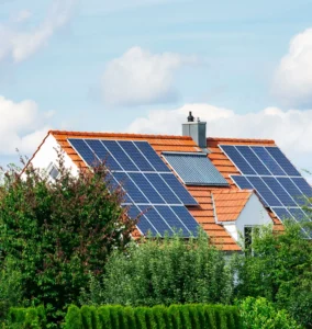 Haus mit rotem Ziegeldach, auf dem mehrere Solarpaneele und ein Solarkollektor installiert sind, umgeben von grünen Bäumen und Sträuchern vor blauem Himmel mit Wolken.