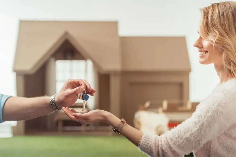 Frau mit hellem Pullover erhält lächelnd einen Schlüssel von einer anderen Person, im Hintergrund unscharf ein Modellhaus, symbolisiert Übergabe oder Immobilienübergabe.