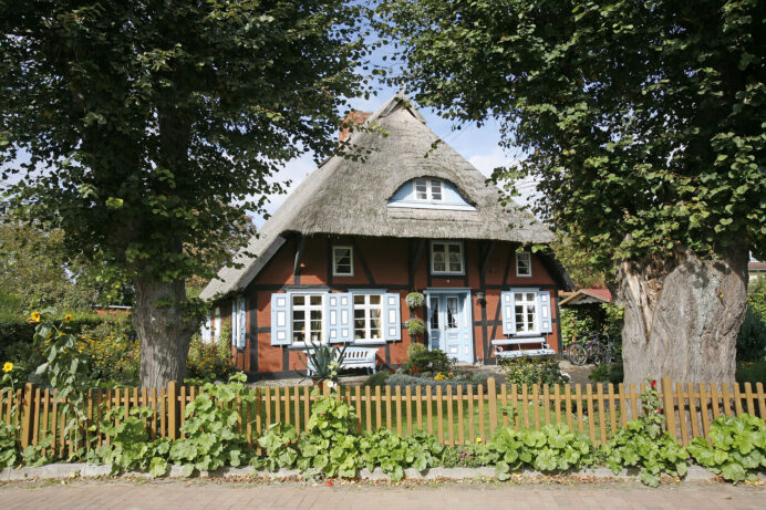 Fachwerkhaus mit Reetdach, roter Fassade und hellblauen Fensterläden, umgeben von einem Garten mit Blumen und zwei großen Bäumen, davor ein niedriger Holzzaun, im Hintergrund Fahrräder und blauer Himmel.