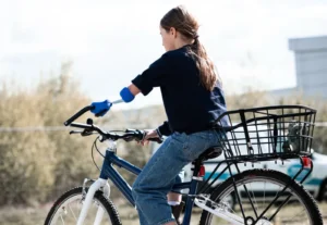 Mädchen mit blauer Prothese am linken Unterarm fährt Fahrrad mit schwarzem Gepäckkorb, trägt dunkelblauen Pullover und Jeans, unscharfer Hintergrund mit Büschen und Gebäuden.