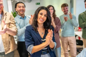 Gruppe von fünf jungen Erwachsenen in einem modernen Büro, die lächelnd und applaudierend zusammenstehen, im Vordergrund eine Frau mit dunklen, lockigen Haaren und blauem Blazer, die direkt in die Kamera lächelt.