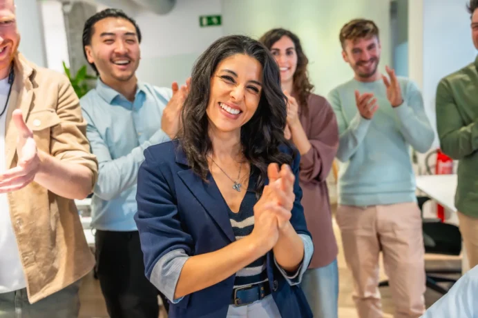 Gruppe von fünf jungen Erwachsenen in einem modernen Büro, die lächelnd und applaudierend zusammenstehen, im Vordergrund eine Frau mit dunklen, lockigen Haaren und blauem Blazer, die direkt in die Kamera lächelt.