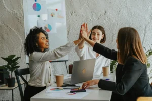 Drei Frauen in einem modernen Büro geben sich lächelnd einen High-Five, sitzen an einem Tisch mit Laptop, Kaffeebechern und Diagrammen, im Hintergrund Whiteboard mit bunten Kreis- und Balkendiagrammen, symbolisiert Teamarbeit und erfolgreiche Zusammenarbeit.