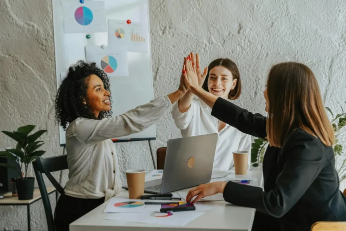 Drei Frauen in einem modernen Büro geben sich lächelnd einen High-Five, sitzen an einem Tisch mit Laptop, Kaffeebechern und Diagrammen, im Hintergrund Whiteboard mit bunten Kreis- und Balkendiagrammen, symbolisiert Teamarbeit und erfolgreiche Zusammenarbeit.