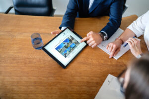 Zwei Personen sitzen an einem Holztisch, eine zeigt auf ein Tablet mit einer Website, die mehrere Bilder von Gebäuden und Text enthält. Neben dem Tablet steht ein Glas Wasser, beide Personen haben Notizblöcke vor sich. Die Szene vermittelt eine Beratung oder Besprechung im professionellen Umfeld.