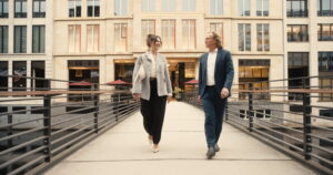 Zwei Personen, eine Frau und ein Mann, gehen auf einer modernen Fußgängerbrücke vor einem Bürogebäude mit großen Fenstern. Die Frau trägt eine helle Bluse, eine graue Jacke, schwarze Hose und helle Schuhe, der Mann einen dunkelblauen Anzug mit weißem Hemd und schwarzen Schuhen. Beide lächeln und blicken sich an.
