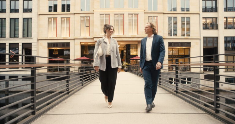 Zwei Personen, eine Frau und ein Mann, gehen auf einer modernen Fußgängerbrücke vor einem Bürogebäude mit großen Fenstern. Die Frau trägt eine helle Bluse, eine graue Jacke, schwarze Hose und helle Schuhe, der Mann einen dunkelblauen Anzug mit weißem Hemd und schwarzen Schuhen. Beide lächeln und blicken sich an.