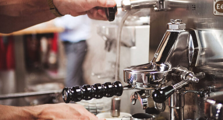 Hände bedienen eine professionelle Espressomaschine aus Edelstahl, eine Hand hält den Siebträger, die andere betätigt den Hebel zum Brühen, im Hintergrund unscharf eine Person und Café-Atmosphäre.