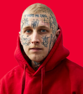 Mann mit zahlreichen schwarzen Gesichtstattoos, trägt roten Kapuzenpullover, roter einfarbiger Hintergrund, Portraitaufnahme.