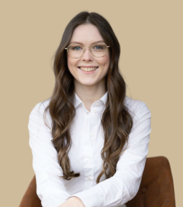Junge Frau mit langen braunen Haaren und Brille, trägt eine weiße Bluse, sitzt vor einfarbig beigem Hintergrund und lächelt freundlich.
