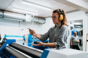 Frau mit Schutzbrille und Gehörschutz bedient in einer Werkstatt eine blaue Maschine, die ein dünnes Metallband formt; im Hintergrund unscharf ein Mann in grauem T-Shirt.
