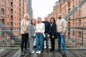 Sechs Personen stehen auf einer Holzbrücke vor historischen roten Backsteingebäuden in Hamburg, lächeln in die Kamera. Sie tragen Business-Casual-Kleidung, darunter Blazer, Jeans und Pullover. Die Szene vermittelt ein professionelles Team in urbaner Umgebung.