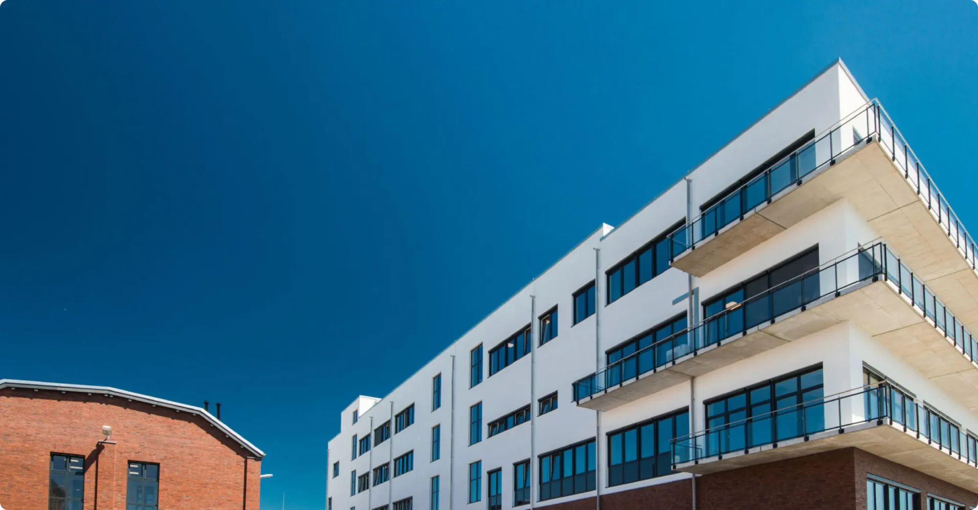 Moderner Bürokomplex mit weißer Fassade, großen Fenstern und Balkonen vor klarem blauem Himmel, links angrenzendes rotes Backsteingebäude, symbolisiert professionelle Unternehmenswebsite und digitales Marketing.