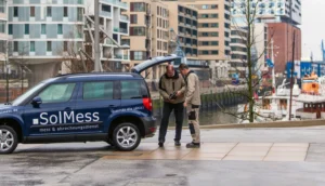 Zwei Männer stehen neben einem blauen Fahrzeug mit der Aufschrift „SolMess mess & abrechnungsdienst“ und „Qualität die zählt!“. Der Kofferraum des Autos ist geöffnet, die Männer betrachten gemeinsam ein Gerät. Im Hintergrund moderne Wohngebäude und ein Hafen mit Booten.