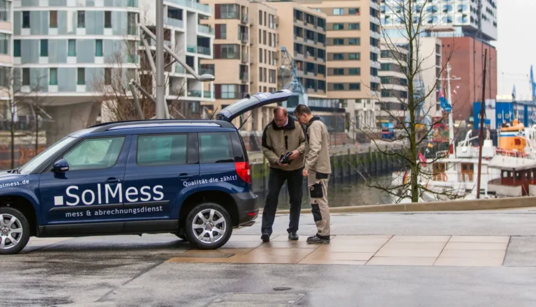 Zwei Männer stehen neben einem blauen Fahrzeug mit der Aufschrift „SolMess mess & abrechnungsdienst“ und „Qualität die zählt!“. Der Kofferraum des Autos ist geöffnet, die Männer betrachten gemeinsam ein Gerät. Im Hintergrund moderne Wohngebäude und ein Hafen mit Booten.