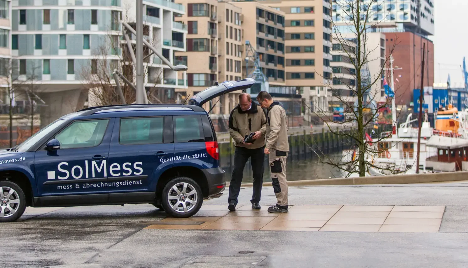 Zwei Männer stehen neben einem blauen Fahrzeug mit der Aufschrift „SolMess mess & abrechnungsdienst“ und „Qualität die zählt!“. Der Kofferraum des Autos ist geöffnet, die Männer betrachten gemeinsam ein Gerät. Im Hintergrund moderne Wohngebäude und ein Hafen mit Booten.