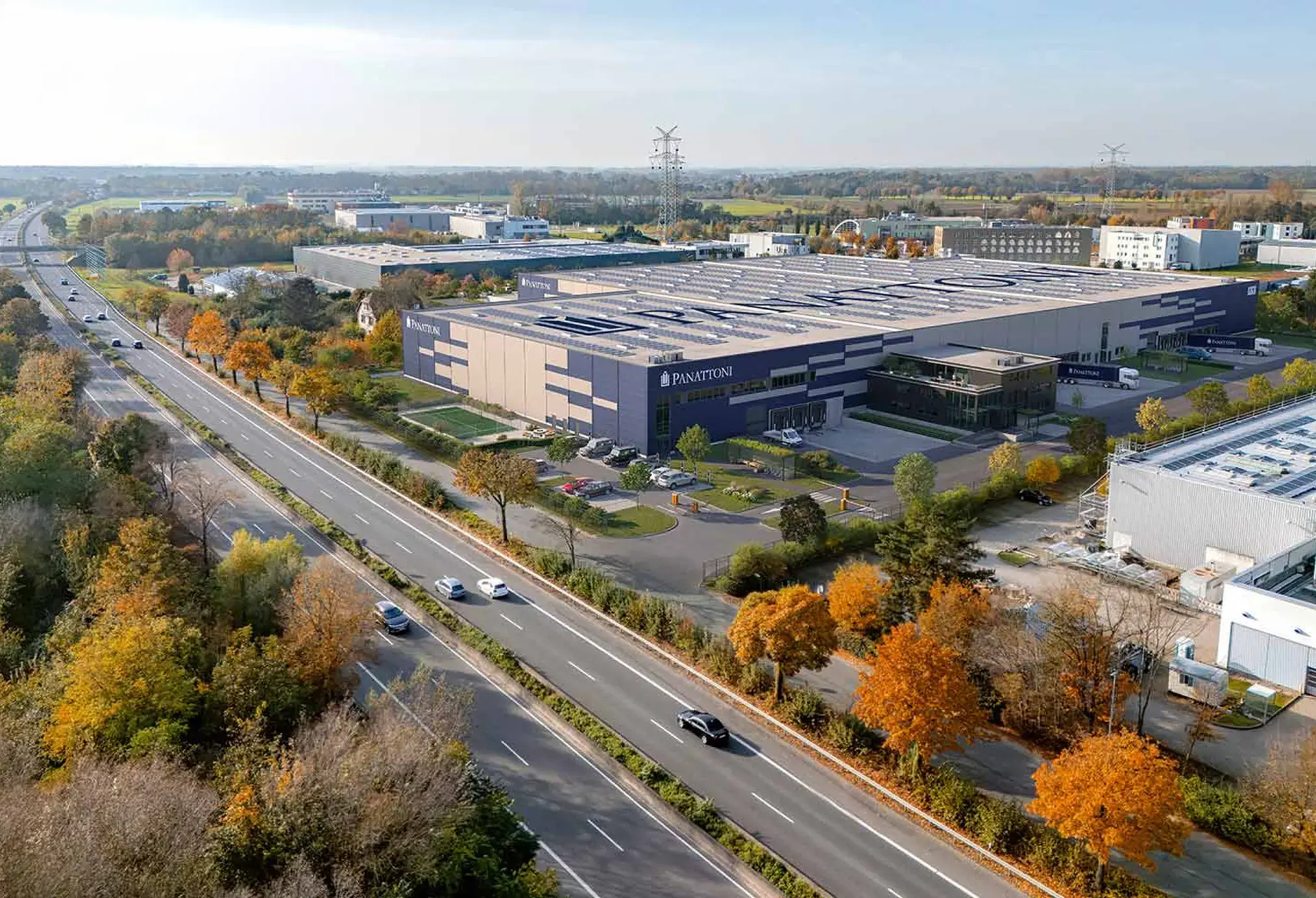 Großes Industrie- und Logistikgebäude mit dem Schriftzug „IBISQUS“ an der Fassade, umgeben von herbstlich gefärbten Bäumen und angrenzend an eine mehrspurige Straße mit mehreren Fahrzeugen, im Hintergrund weitere Gewerbegebäude und flaches Landschaftsgebiet.