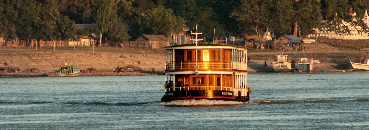 Dreistöckiges, traditionell gestaltetes Passagierschiff auf einem breiten Fluss, im Hintergrund Ufer mit Bäumen, kleinen Gebäuden und weiteren Booten, Sonnenlicht spiegelt sich auf der Holzverkleidung des Schiffes.