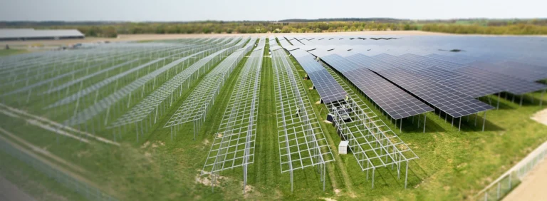Großflächige Freiland-Solaranlage im Bau mit teilweise montierten Solarmodulen auf Metallgestellen, grüne Wiese und weiter Horizont im Hintergrund.