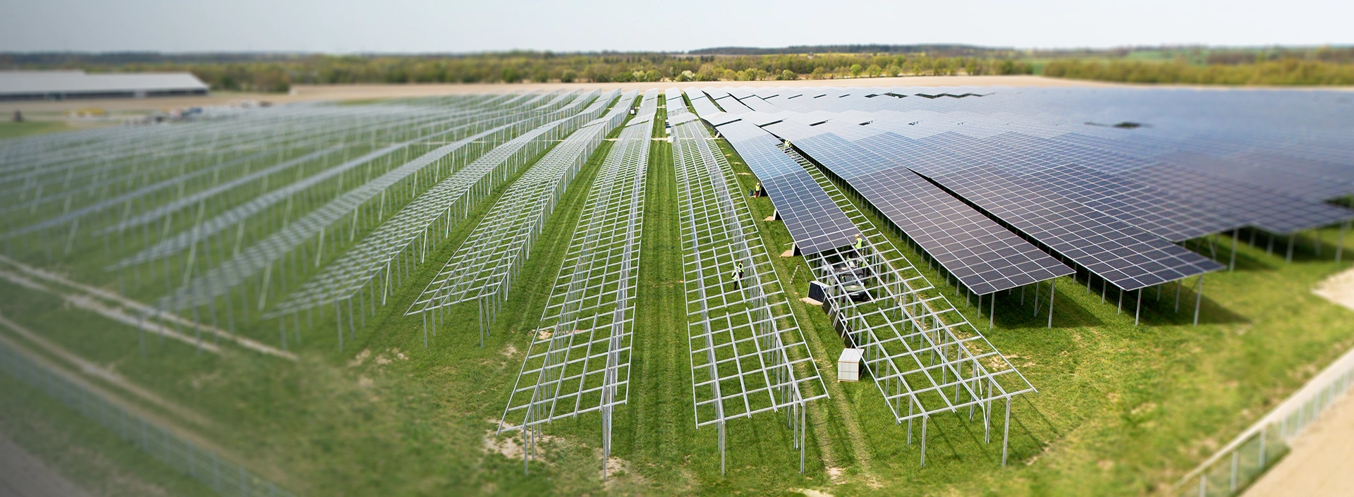 Großflächige Freiland-Solaranlage im Bau mit teilweise montierten Solarmodulen auf Metallgestellen, grüne Wiese und weiter Horizont im Hintergrund.
